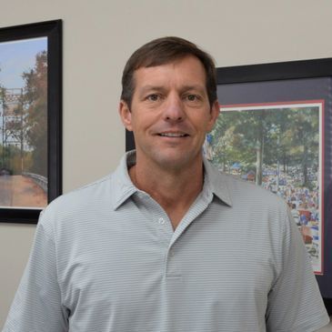 Man in a gray polo shirt smiles, standing in front of framed artwork.