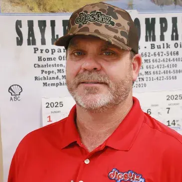Man in red polo shirt and camouflage hat in front of a calendar and business signage.