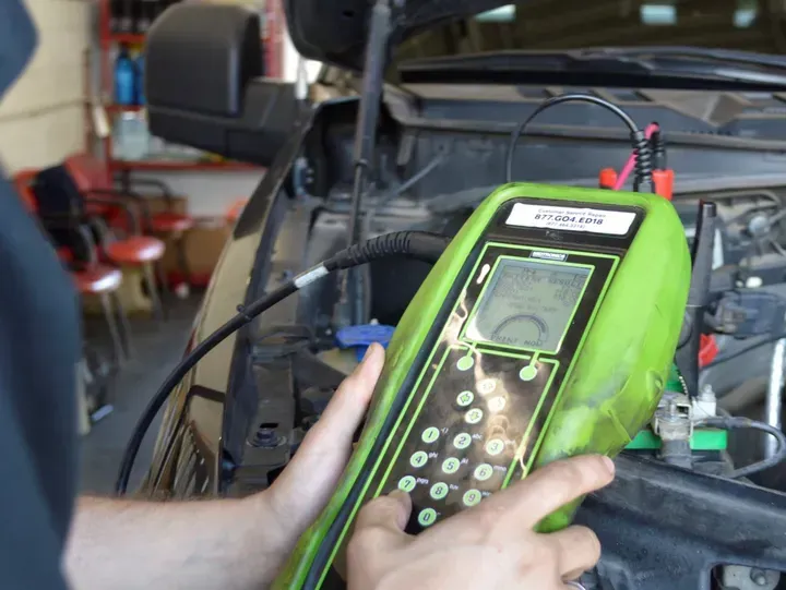 Mechanic using a green battery tester on a black vehicle's open hood in a garage.
