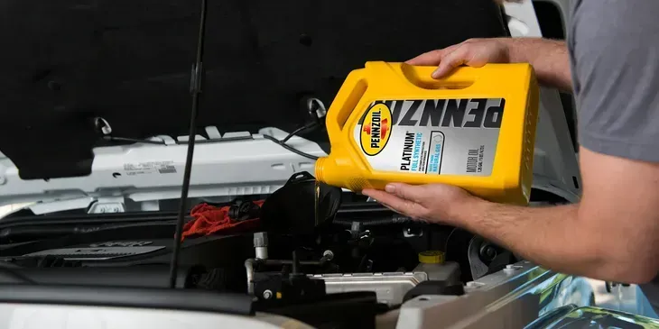 Person adding motor oil to a car engine. They hold a yellow Pennzoil bottle over the open hood.