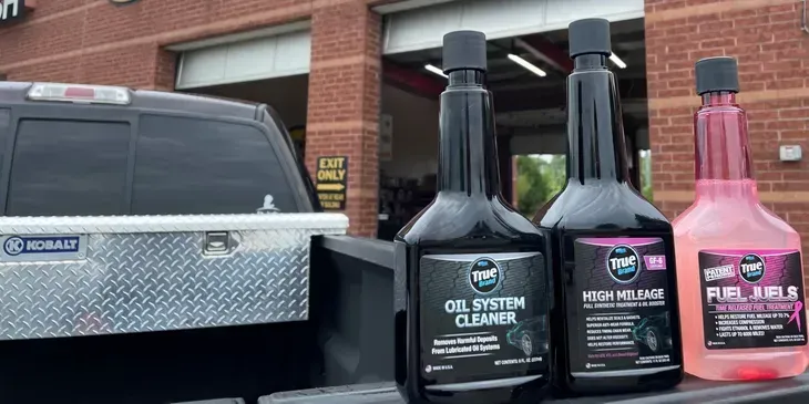 Three bottles of car additives on a truck bed, in front of a brick building and garage.