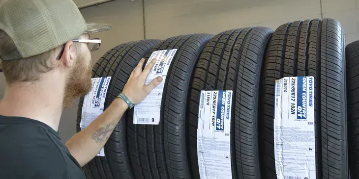 Man examines tire options in a shop, hand on a tire. Dark tires, white labels, and a cap.