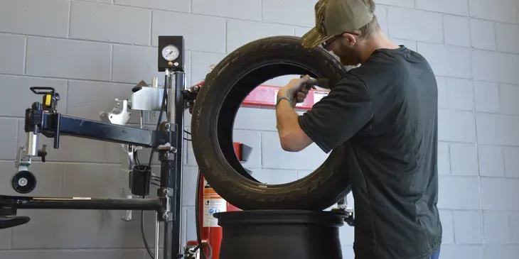 A mechanic working on a tire at a tire shop. He wears a hat and is using a tool to fix the tire.