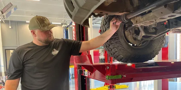 Man in camo hat checks underside of vehicle on a lift in a shop.