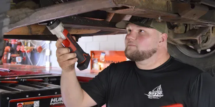 A man wearing a baseball cap, uses a flashlight to look under a vehicle.