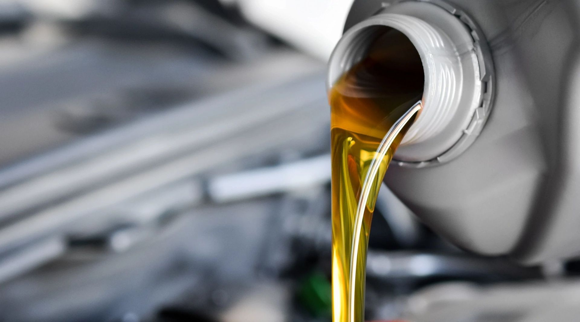 Pouring oil from a gray container into a car engine, showcasing yellow liquid and a blurred background.