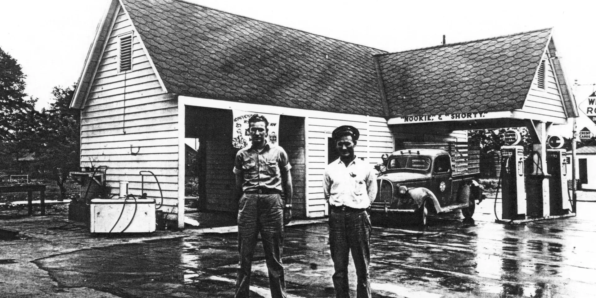 Two men stand outside a vintage gas station with a truck parked nearby.