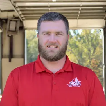 Man with a beard wearing a red polo shirt smiles outdoors; logo on shirt.