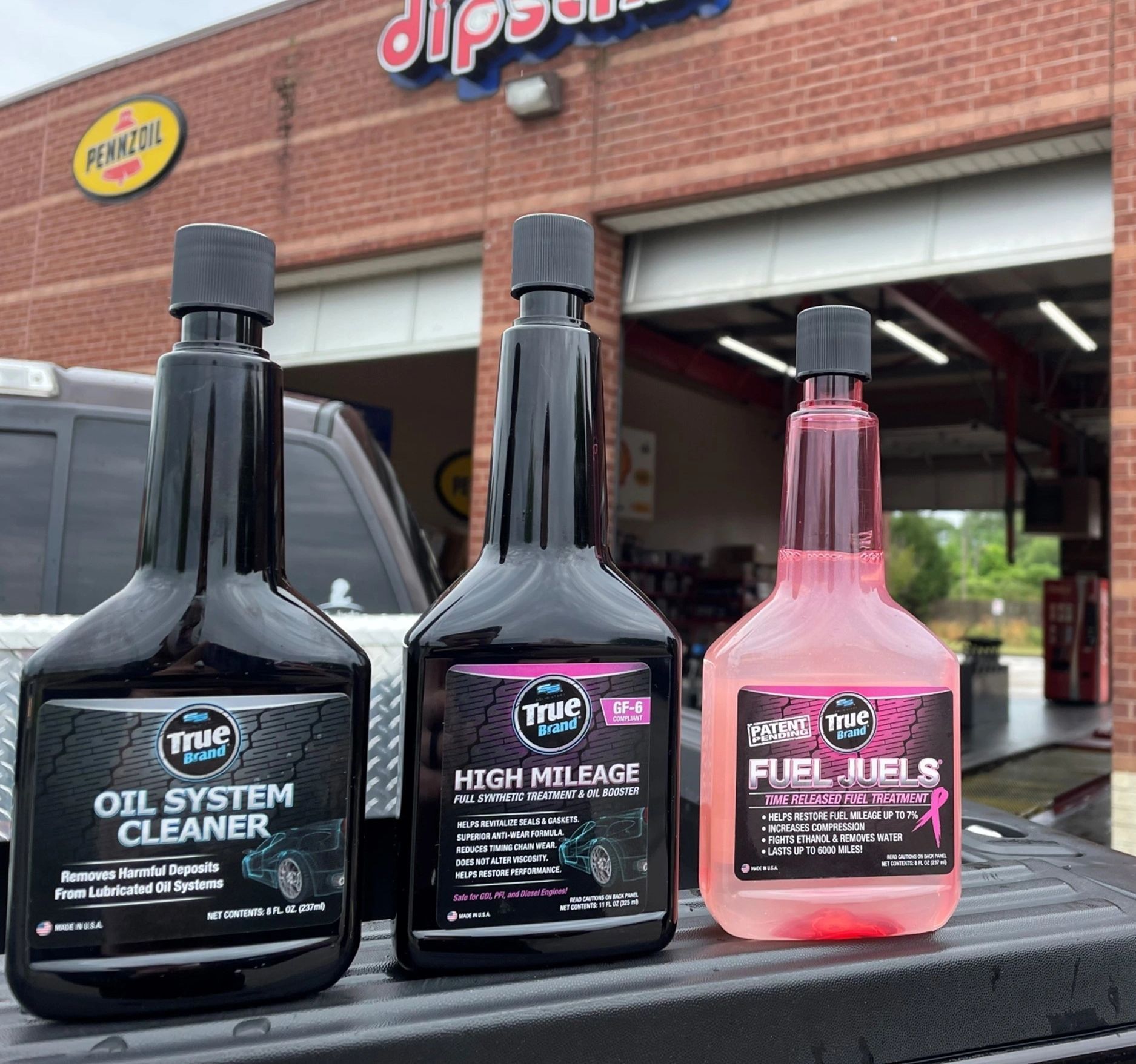 Three fuel additives in bottles on a truck bed: black oil cleaner, high mileage, and pink fuel treatment.