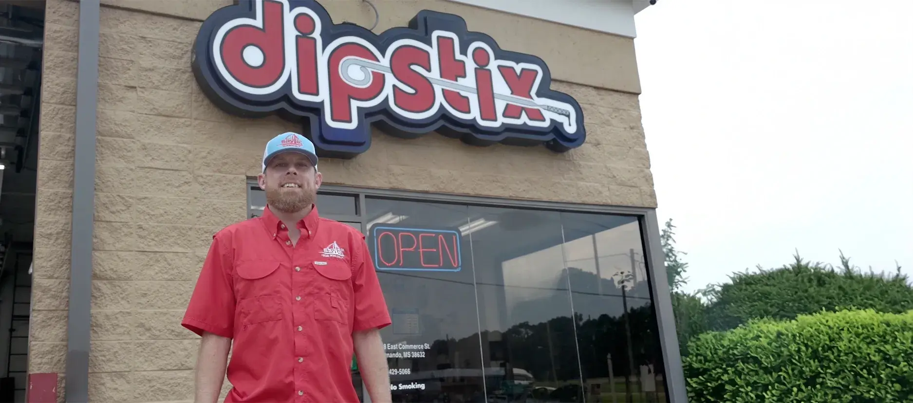 Man in red shirt stands in front of a Dipstix restaurant, open sign in window.