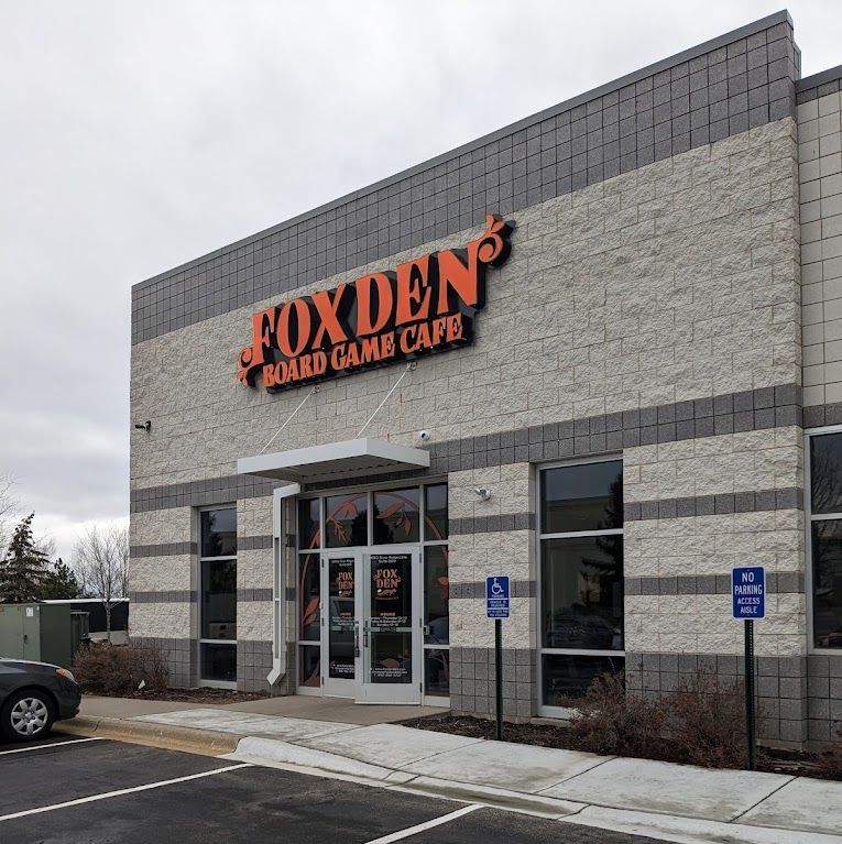 Exterior of Fox Den Board Game Cafe with orange sign on a brick building, accessible parking signs.
