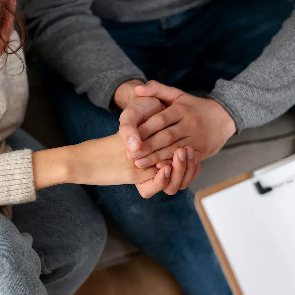 Two people holding hands, one wearing a gray sweater, and a clipboard visible on the right.
