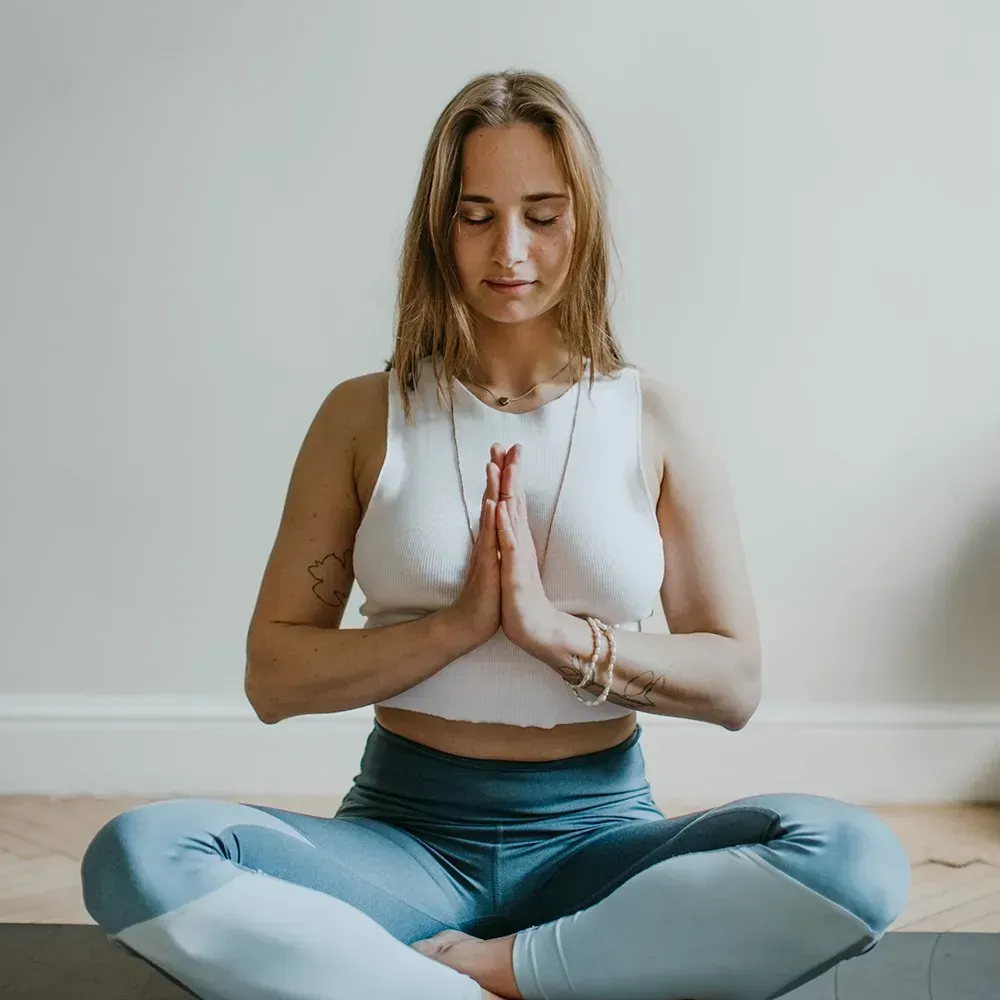 Woman in yoga pose, hands together, eyes closed. Wearing white top and blue leggings, meditating indoors.