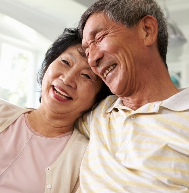 Smiling couple, woman leans head on man’s shoulder indoors. Man wears yellow striped shirt, woman pink top.