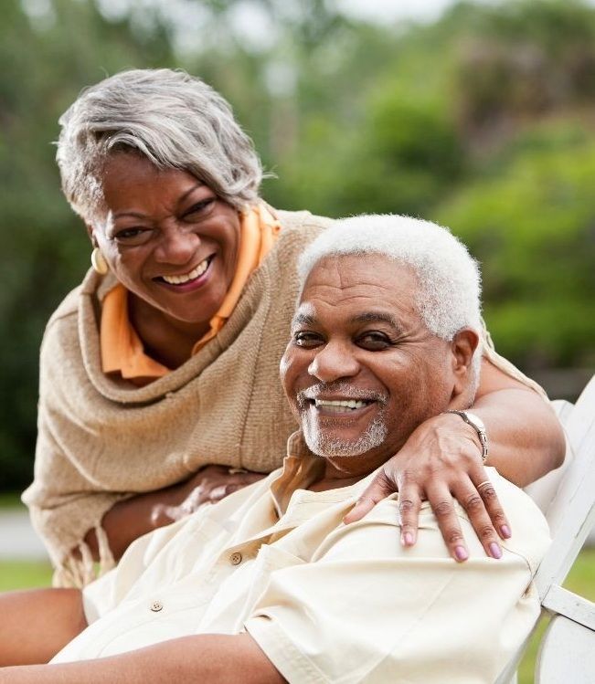 Smiling couple outdoors; woman's arm around man's shoulders. Sunlight, trees in background.