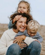 Woman smiling, hugging two children. Blonde girl’s hands on woman's head. Boy hugs woman’s torso. Outdoors.