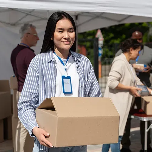 Woman with a blue ID tag holds a cardboard box, volunteering at an outdoor event with other people.