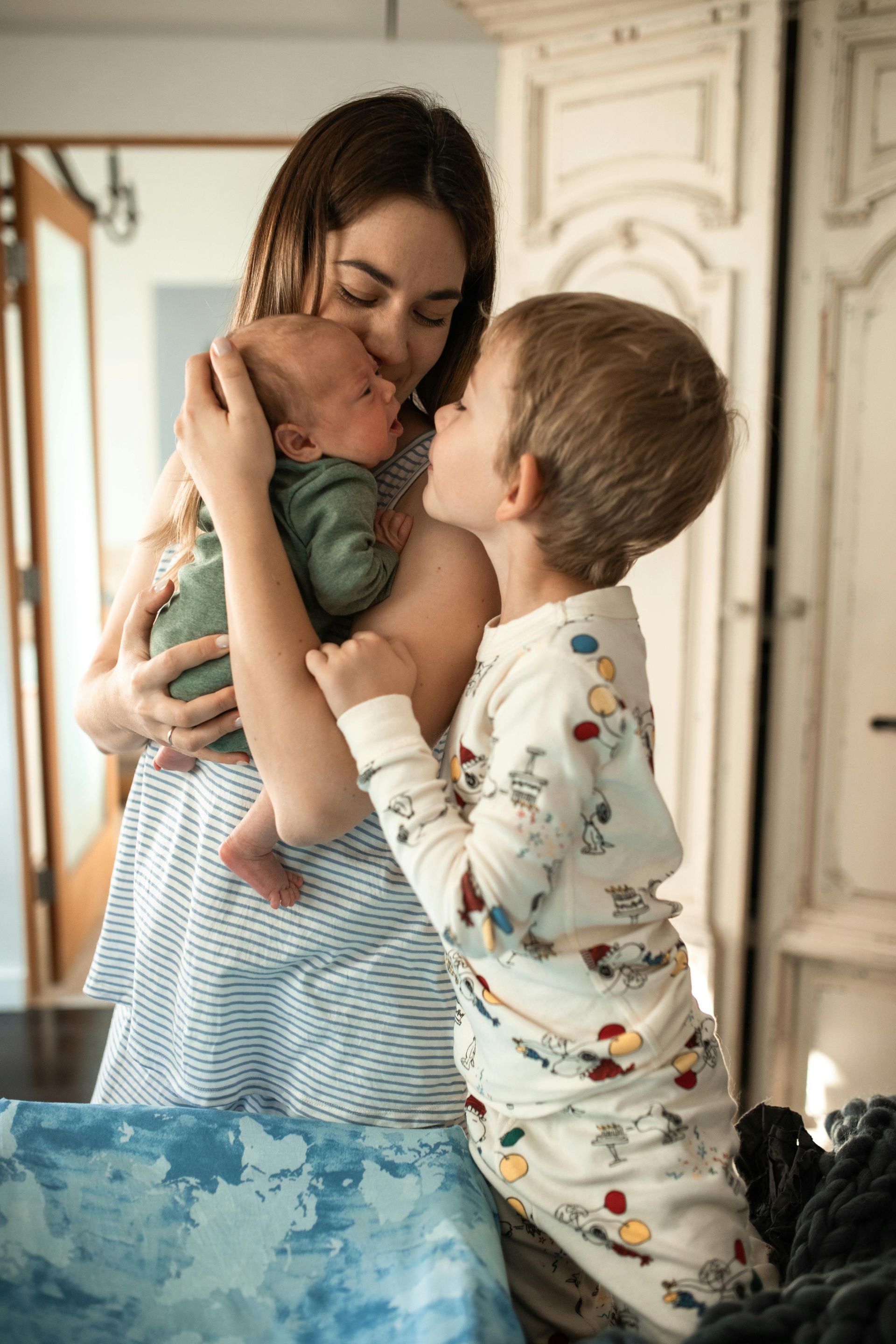 Woman smiling, hugging two children. Blonde girl’s hands on woman's head. Boy hugs woman’s torso. Outdoors.