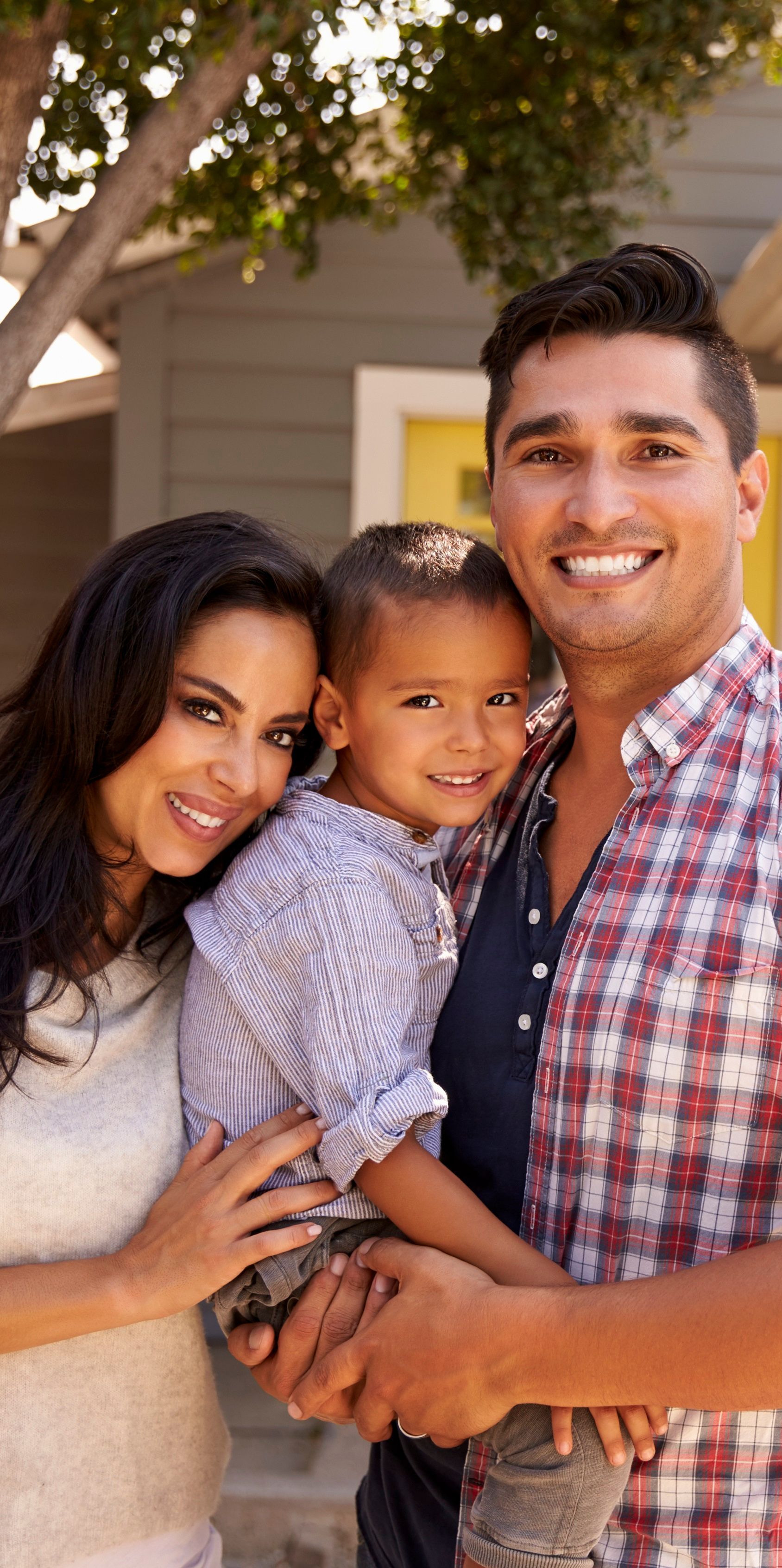 Family of three smiling in front of a house. The man holds the child; the woman embraces them.