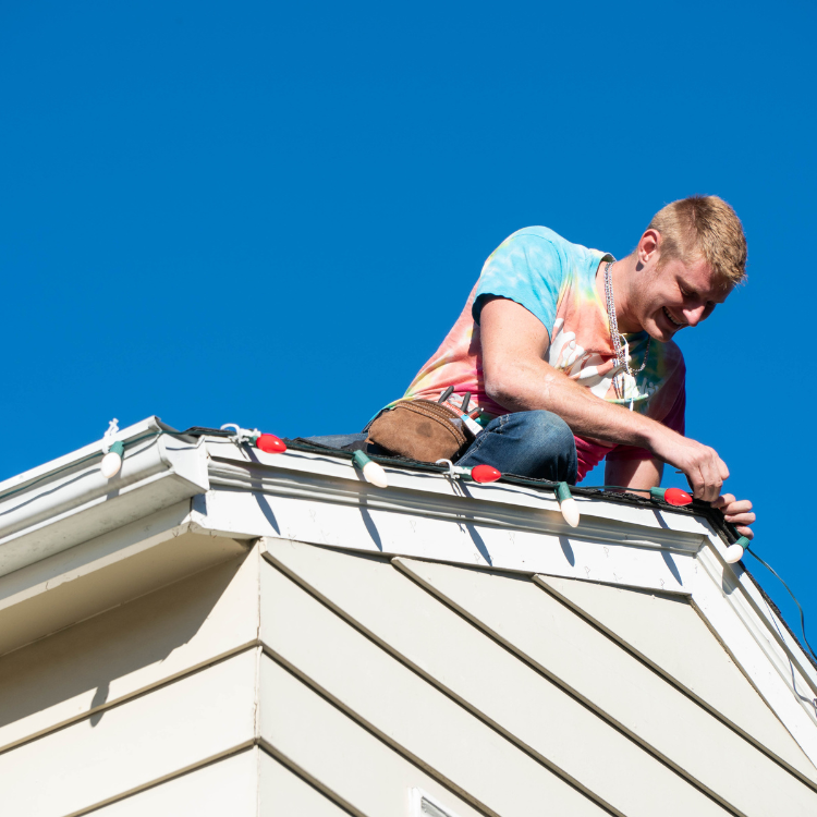 A man is sitting on the roof of a house working on christmas lights