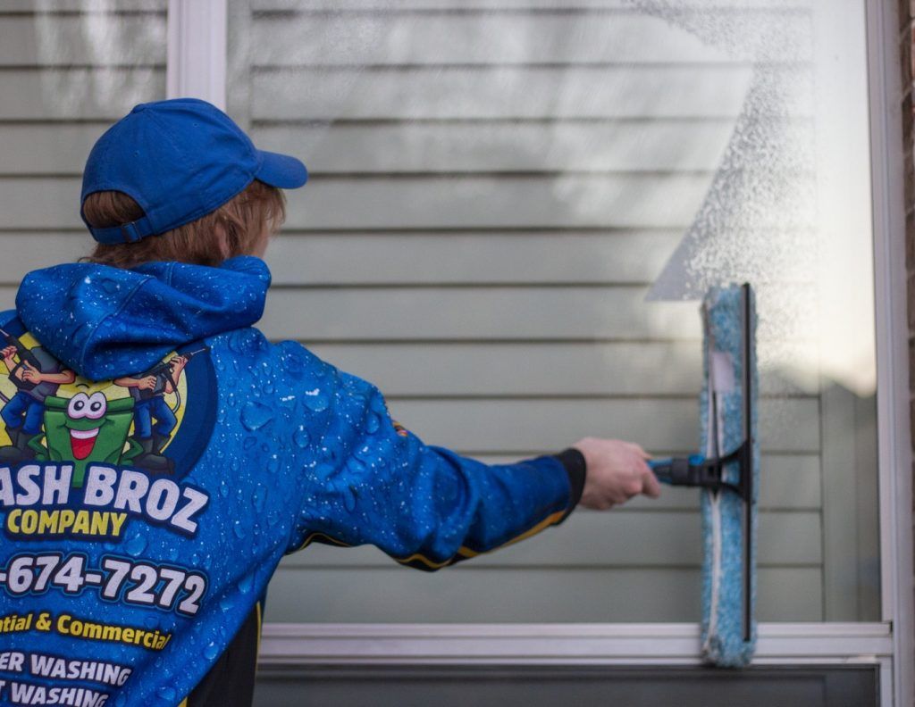 A man wearing a blue jacket that says ash broz company is cleaning a window