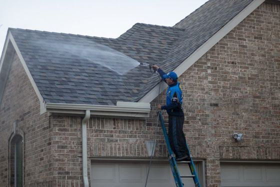 A man on a ladder is cleaning the roof of a brick house.