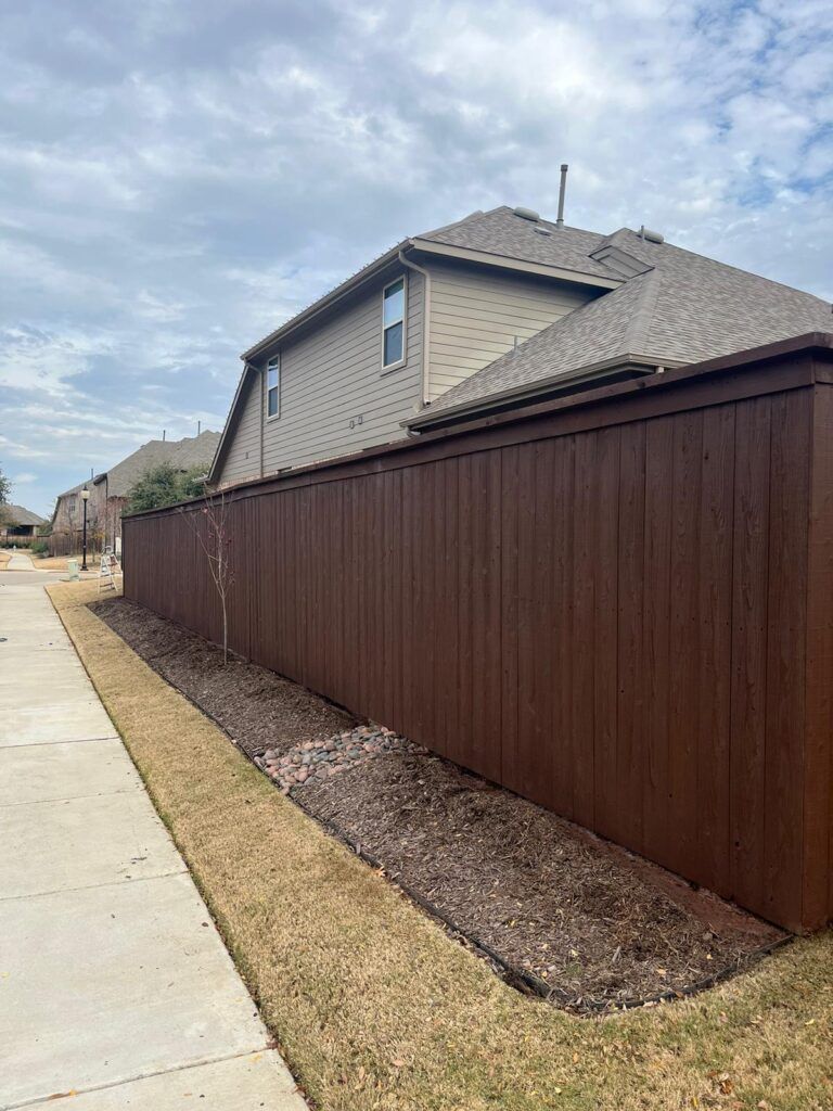 A brown wooden fence is sitting next to a sidewalk in front of a house.