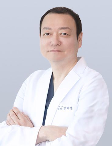 A medical professional in a white coat and dark shirt, standing with arms crossed against a plain, light blue background.