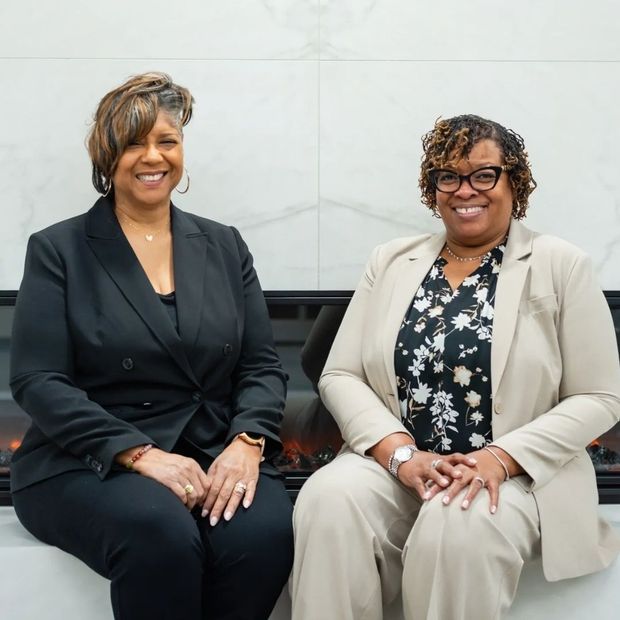 Two women in suits are sitting next to each other and smiling