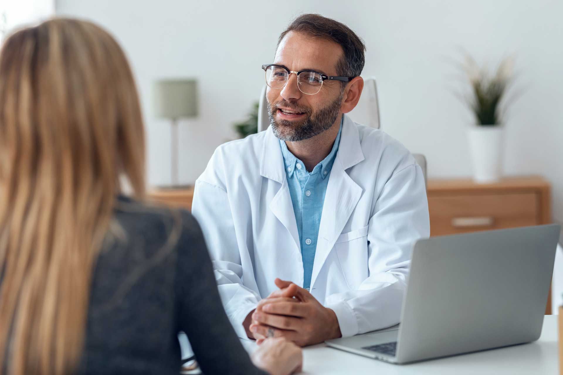 Doctor in white coat speaking with a patient at a desk, laptop present.