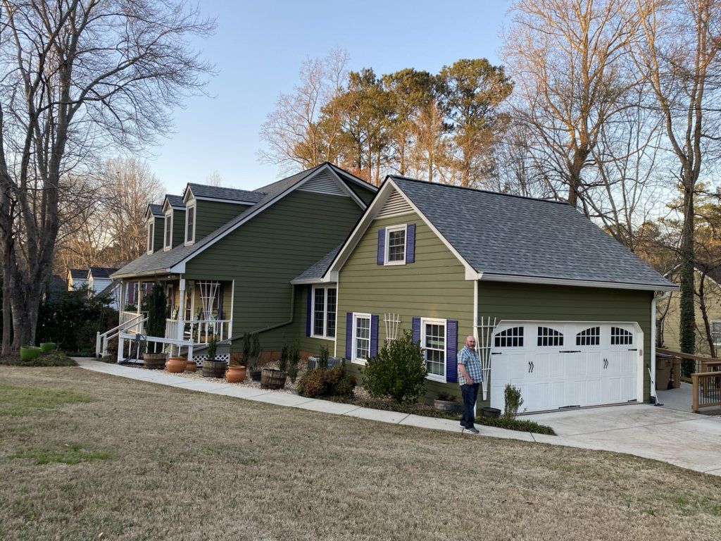 A man is standing in front of a green house with a garage.