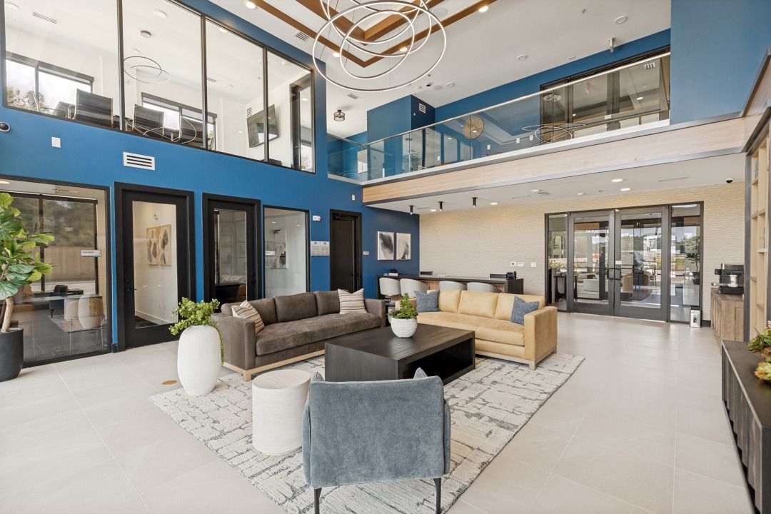 Modern two-story lobby with blue walls, glass railings, and gray seating under a chandelier