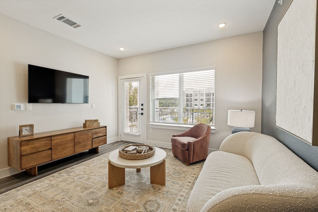 Bright living room with a wall-mounted TV, wooden console, beige sofa, chair, and large window.
