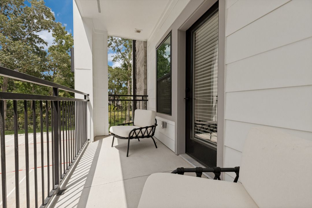 Narrow apartment balcony with black railing, white walls, and two chairs overlooking trees