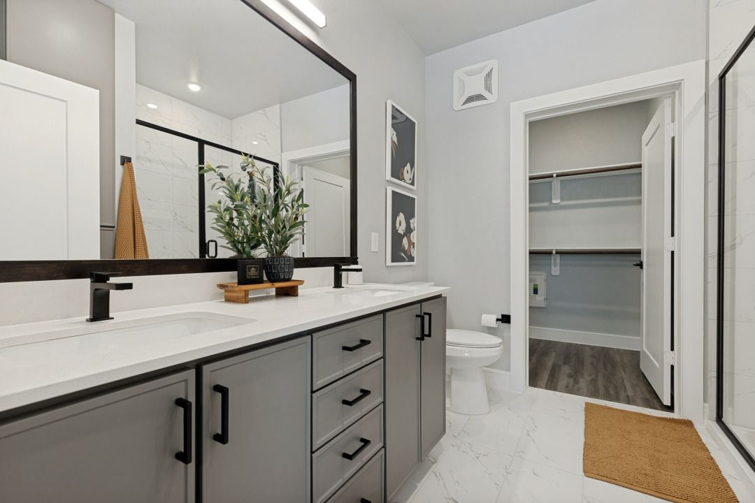 Modern bathroom vanity with gray cabinets, white countertop, large mirror, and walk-in closet beyond.