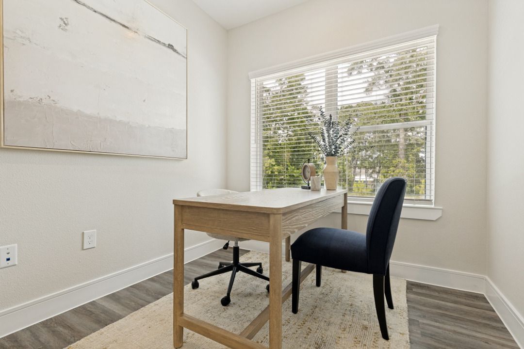 Small home office with a wooden desk, black chair, wall art, and bright window.