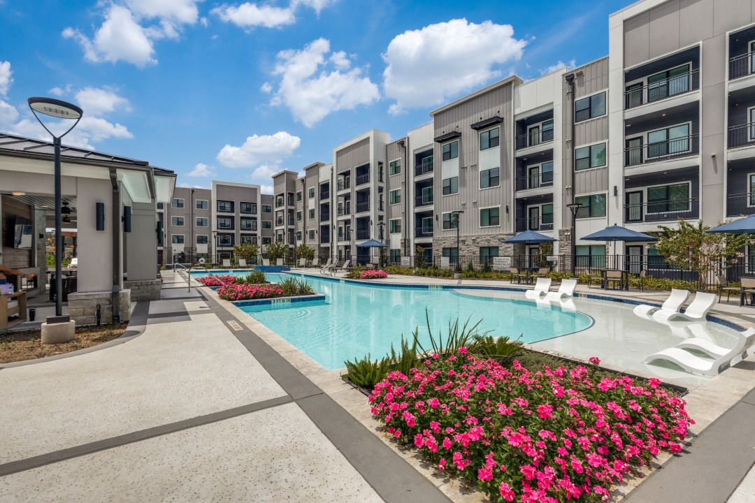 Modern apartment courtyard with a turquoise pool, lounge chairs, and pink flowers under a blue sky