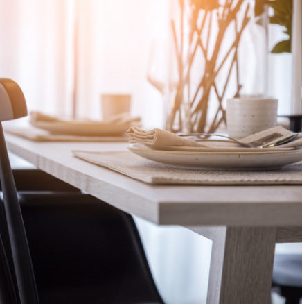 Dining table set for two with plates, napkins, and silverware. Sunlight streams in from a window.