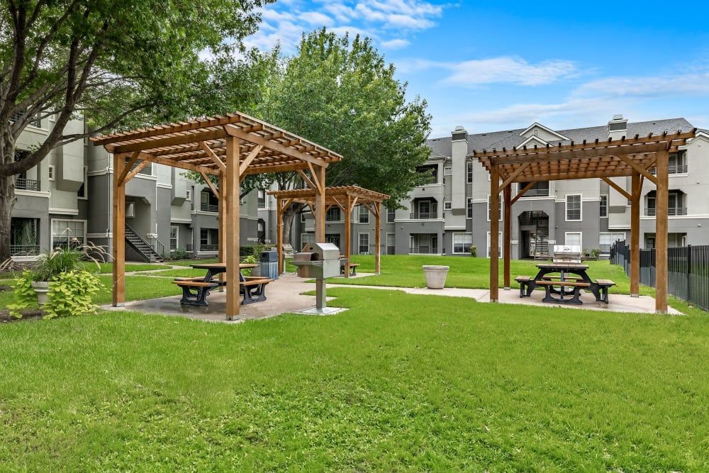A picnic area with a pergola and picnic tables in front of the apartment building at Marquis at Stonegate offers apartments near White Settlement, TX.