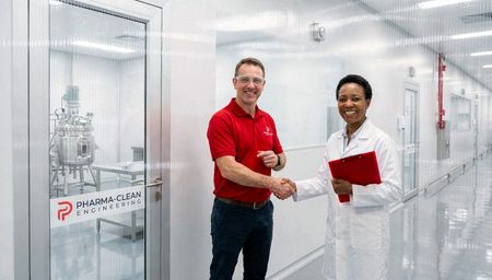 Dois funcionários de laboratório, trajando jalecos brancos e luvas, apertam as mãos em um corredor bem iluminado de uma sala limpa.