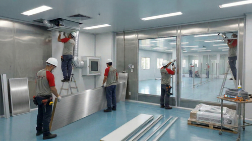 Operários instalando painéis em uma sala moderna e iluminada, com escadas e ferramentas.