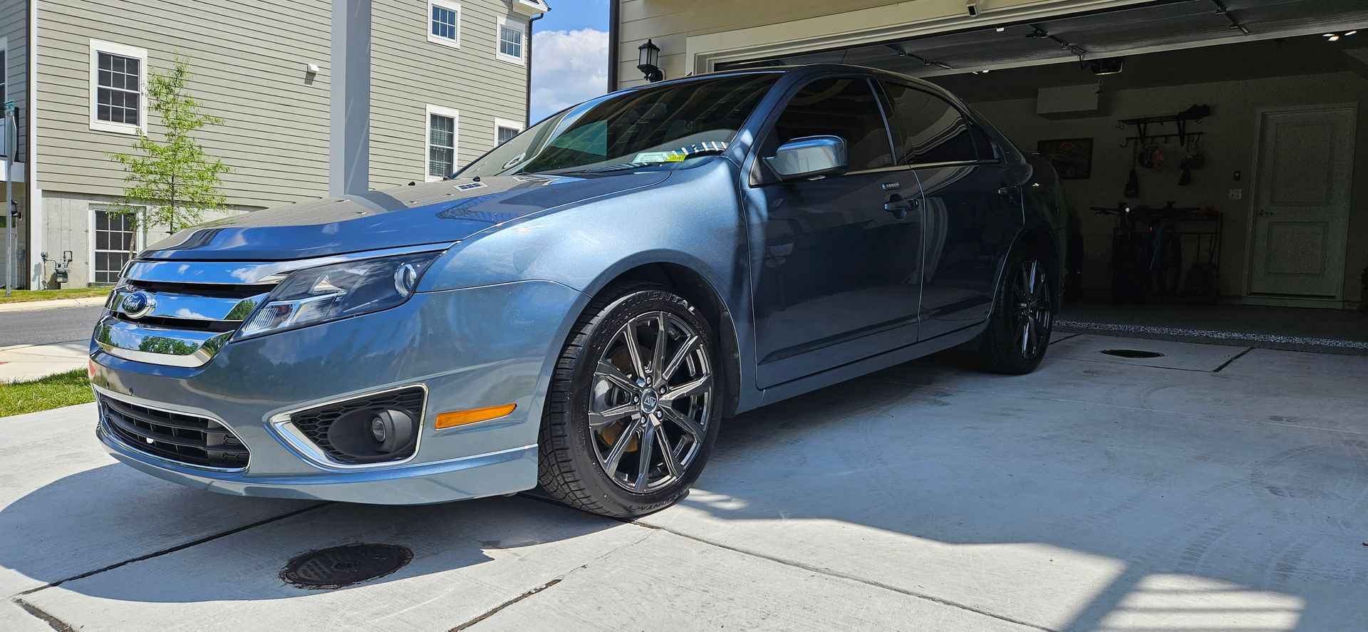 Blue Ford Fusion parked in front of a garage, with buildings in the background under a sunny sky.