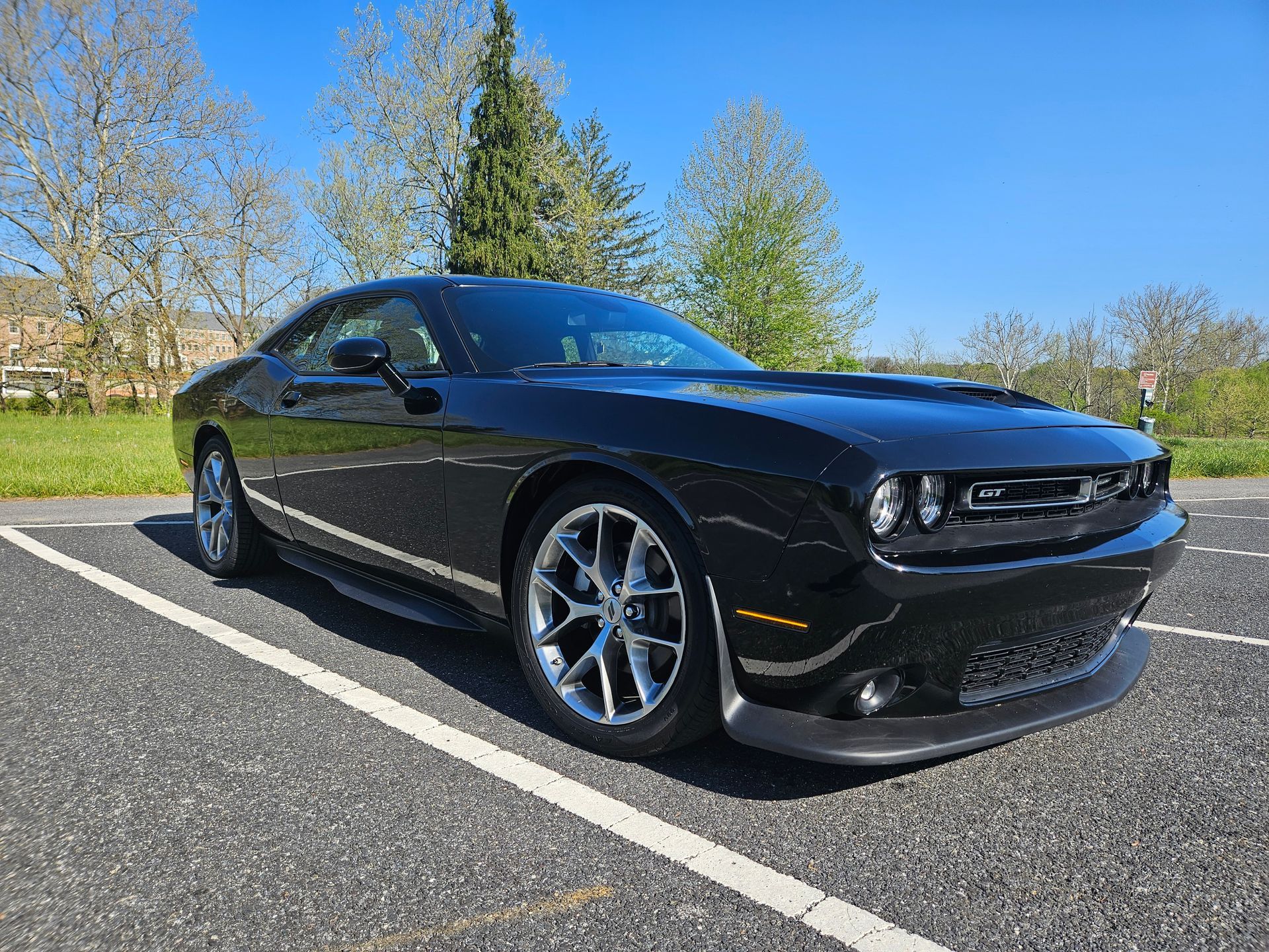 Black Dodge Challenger parked on asphalt, blue sky overhead, with grass and trees in the background.