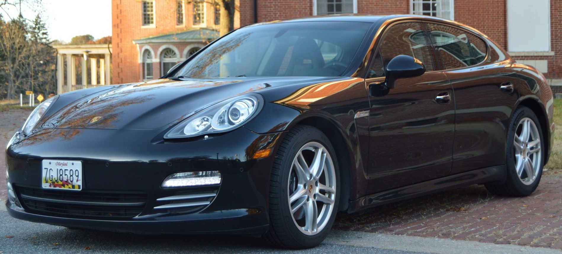Black Porsche Panamera parked on a paved road in front of a brick building with windows.