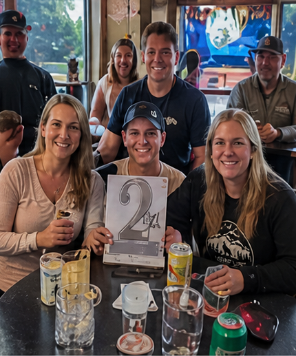 Group seated at a restaurant table, smiling and holding a large “2” sign, with drinks in front.