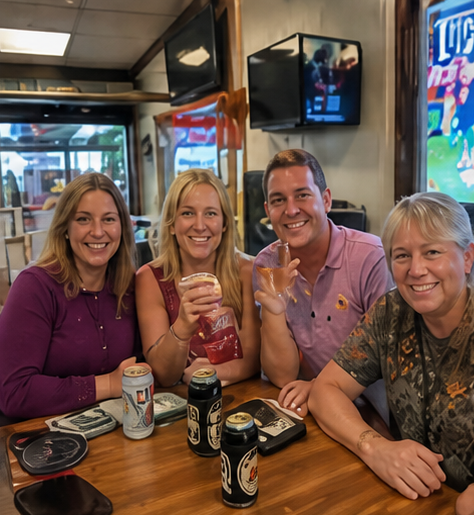 Four friends smiling at a bar table, holding drinks with beer cans and TVs in the background.