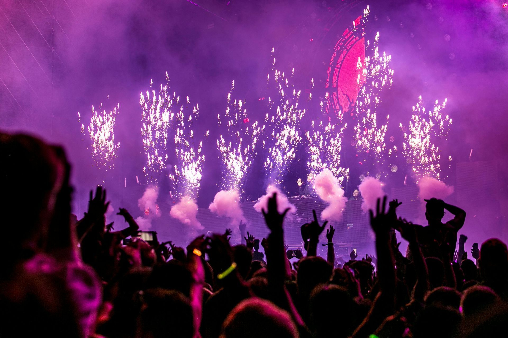 A vibrant concert scene with silhouettes of people cheering amidst purple stage lights and vertical pyrotechnics.