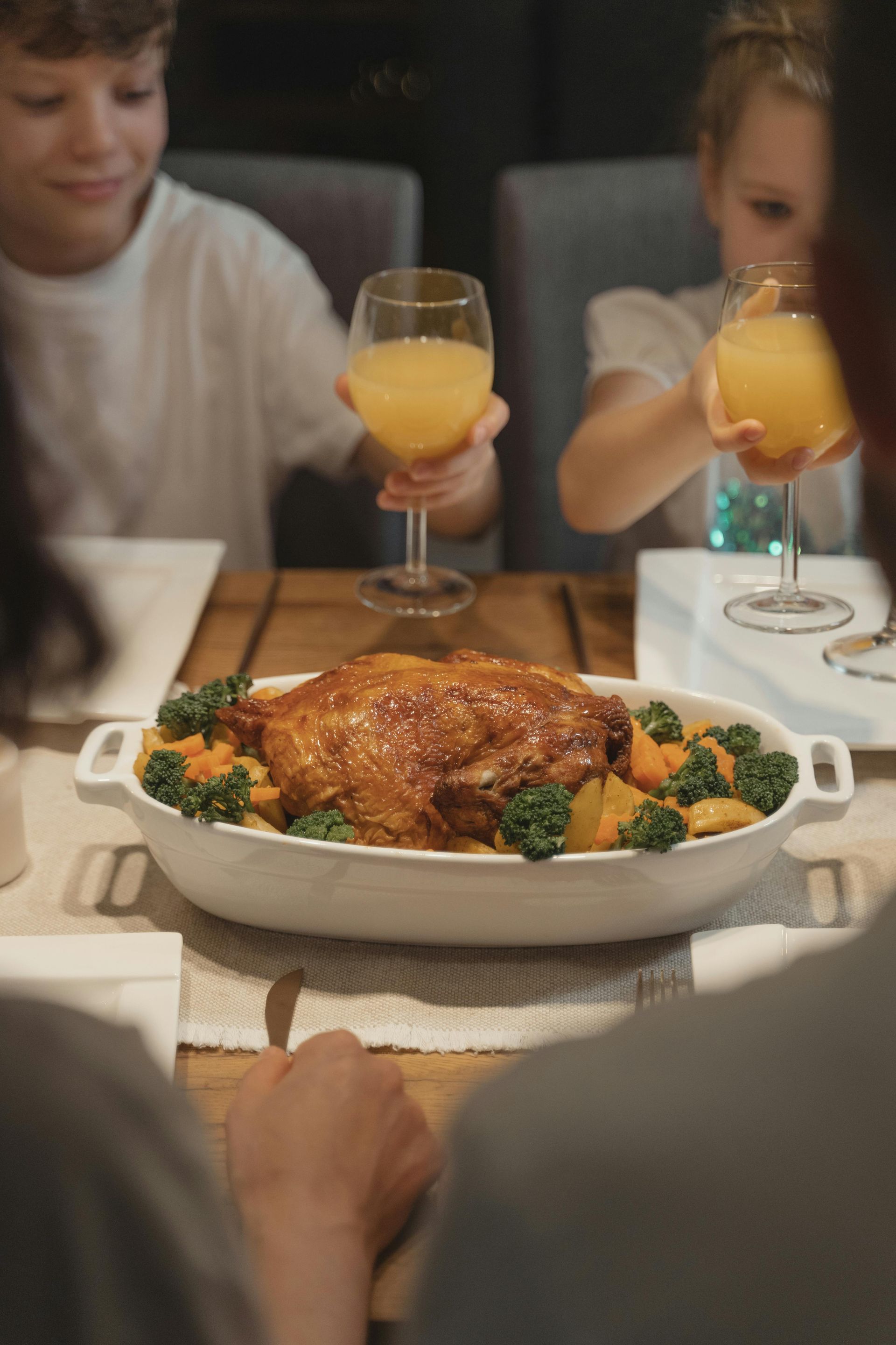 A group of people sitting at a dining table holding glasses of orange juice near a dish of roast chicken with vegetables.