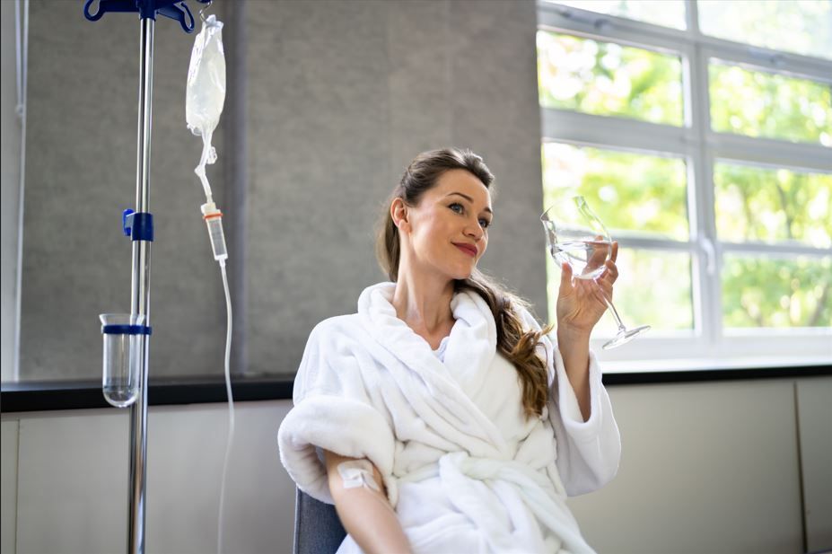 Woman in a white robe receiving an IV drip, holding a glass. Near a window with sunlight.