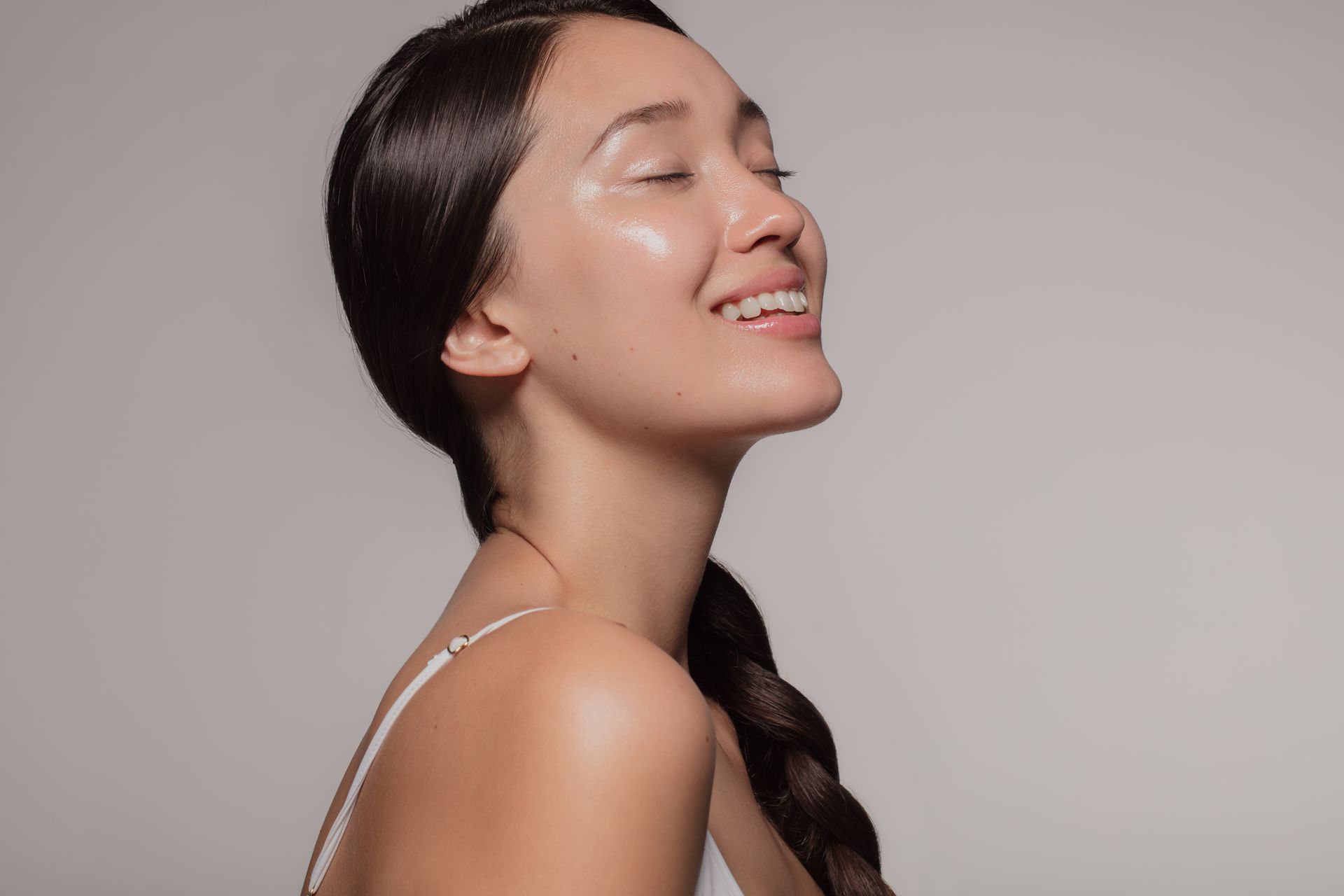 Woman with dark hair in a braid, smiling, looking upwards, glowing skin, wearing a white top, against a gray background.